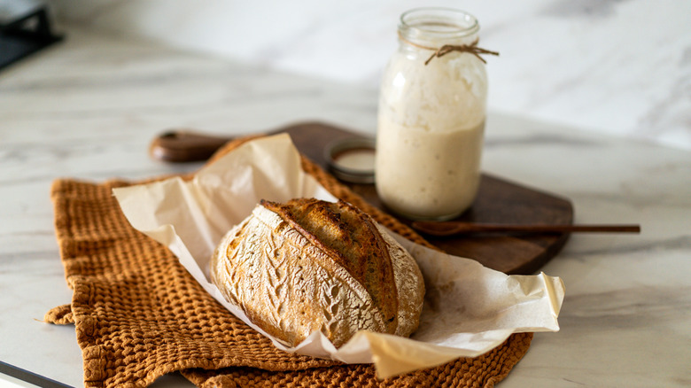 A freshly baked sourdough bread on parchment paper on cloth napkin with raw dough in open glass jar, while an open jar of sourdough starter rests on the background