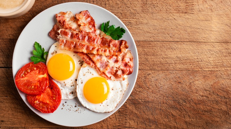 a plate on a wooden table with two fried eggs, bacon and a grilled tomato