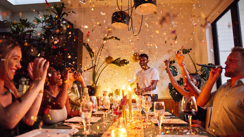 A group of friends gathered around the dinner table as confetti pours down.