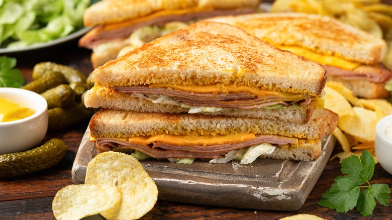 A fried bologna sandwich with cheddar cheese and chopped lettuce on a cutting board, surrounded by cornichons, potato chips, mustard, and lettuce