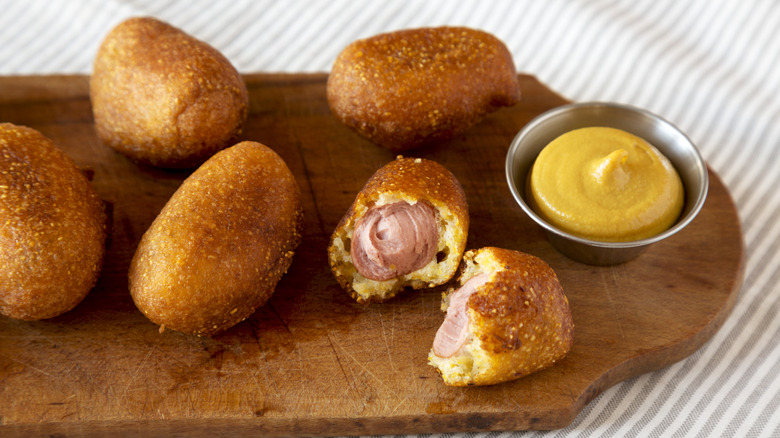 A wooden cutting board with several mini corn dogs and a dipping cup filled with mustard.