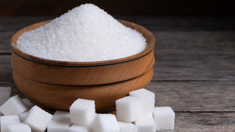 Wooden bowl of sugar next to sugar cubes on wood table.