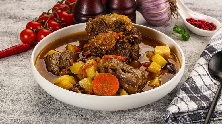 A bowl of oxtail stew with potatoes and carrots, surrounded by a napkin and spoon, with chili flakes, fresh tomatoes, and whole garlic in the background.