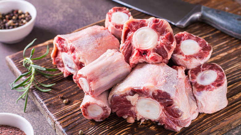 Sliced raw oxtail on a wooden cutting board, a knife to the side, and sprig of rosemary to the other side, and a ramekin of peppercorns in the background.