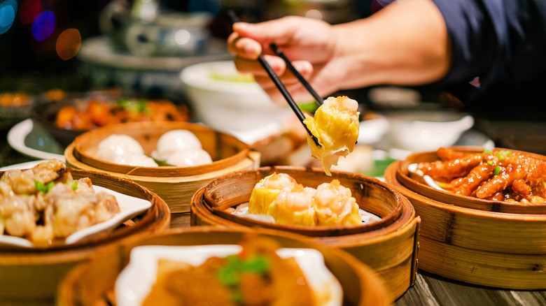 A diner using choptsticks to grab a shumai from a bamboo steamer, with other bamboo steamers filled with food
