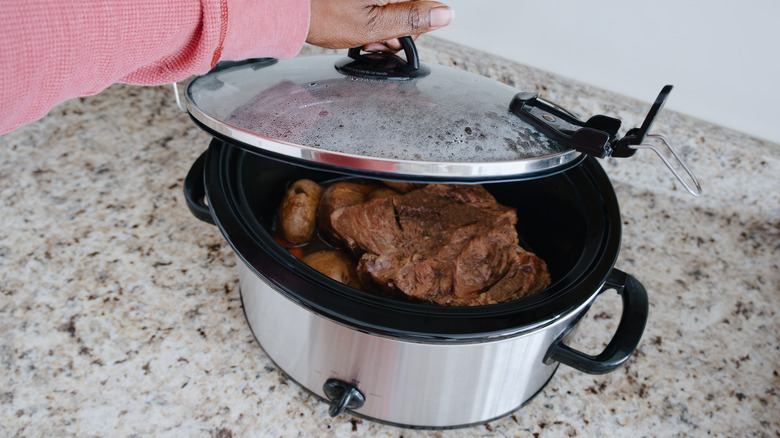 A hand opening the lid of a slow cooker to check the beef cooking within.