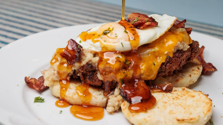 Hot honey being poured over biscuits topped with fried chicken, a fried egg, cheddar, sausage gravy, candy onions, and bacon