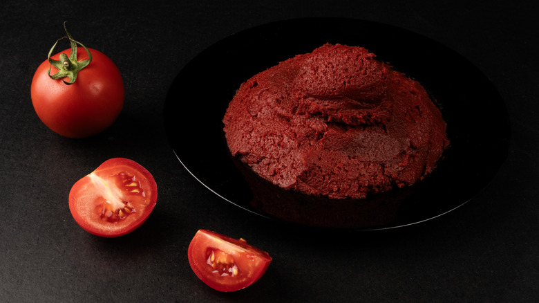 Tomato paste on a dark plate next to a whole tomato and tomato slices