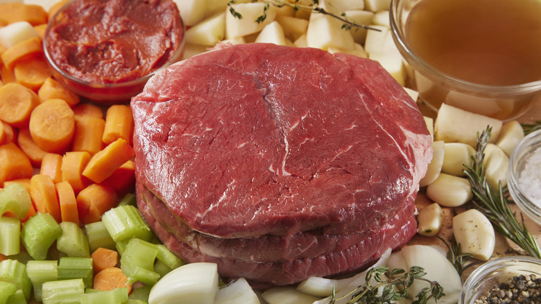 An uncooked cut of beef placed atop chopped carrots, celery, and garlic cloves