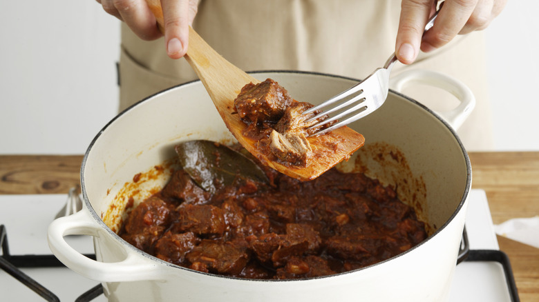 Hands holding wooden spoon and fork pull pieces of cooked beef from pot of stew