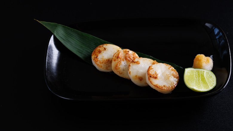 three grilled Hokkaido scallops on a black plate with a leaf and slice of lemon on a black background