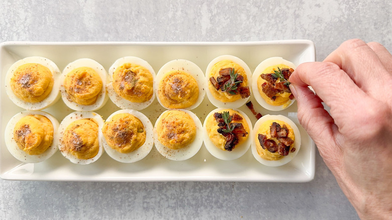 Topping deviled eggs with bacon and thyme sprigs, nutmeg and sea salt