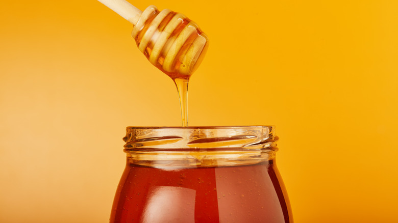 A honey dipper dripping a line of honey into a jar of honey on an orange background.