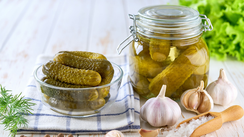 A bowl full of homemade pickles on a tea towel, next to a full jar of pickles, with cloves of garlic and salt in front.