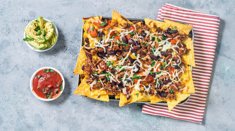 an overhead shot of nachos topped with chili con carne on top of a grey table with a red striped dish towel and two small rameskins filled with avocado slices and salsa