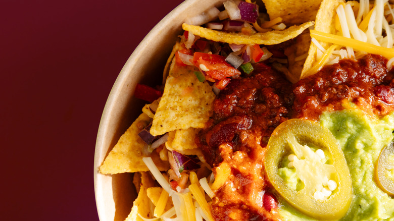 Top view of a plate of nachos on a red table in a white bowl with jalapenos visible