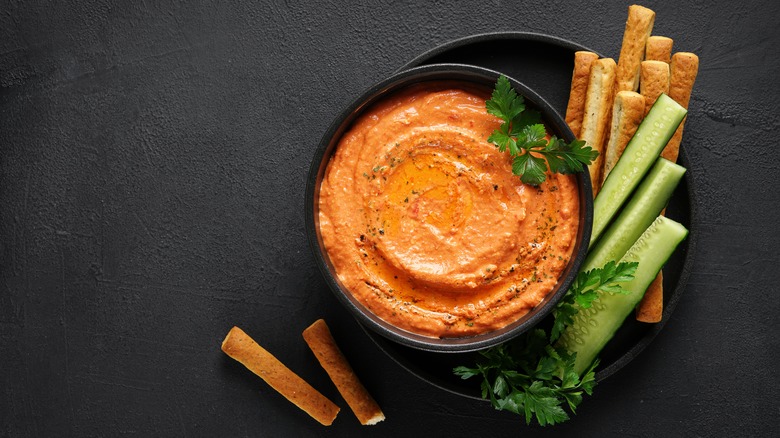 red pepper and feta dip in black bowl with bread sticks and cucumbers, on dark background. top view,