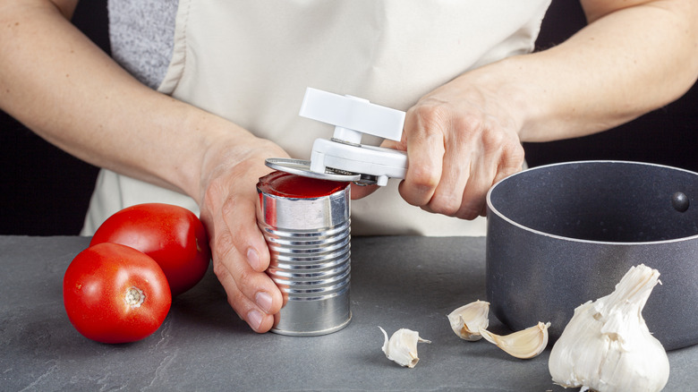 A person is carefully opening a can of tomato paste on a kitchen counter using a white plastic can opener. She is preparing a meal for which she uses both fresh and pureed preserved tomatoes