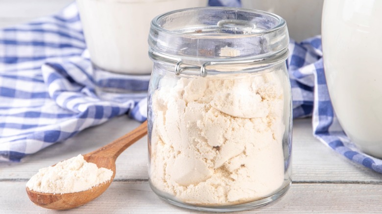 A glass jar of buttermilk powder next to wood spoon with buttermilk powder on wood surface with blue and white checkered cloth