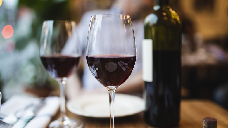 Two wine glasses half-filled with red wine sitting on a tabletop with a plate, napkin, silverware, and a bottle of red wine in the background.