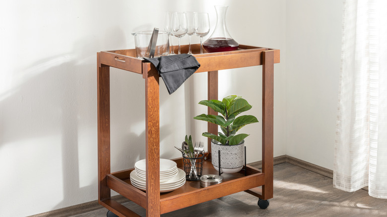 A wooden bar cart holds a carafe of red wine and glasses on its top shelf and white plates, silverware, and a plant on the lower level
