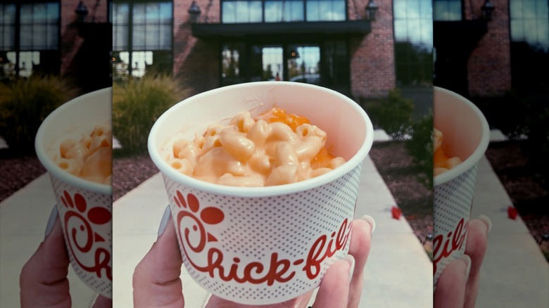 Hand holding up a cup of Chick-fil-A mac and cheese in front of a Chick-fil-A restaurant