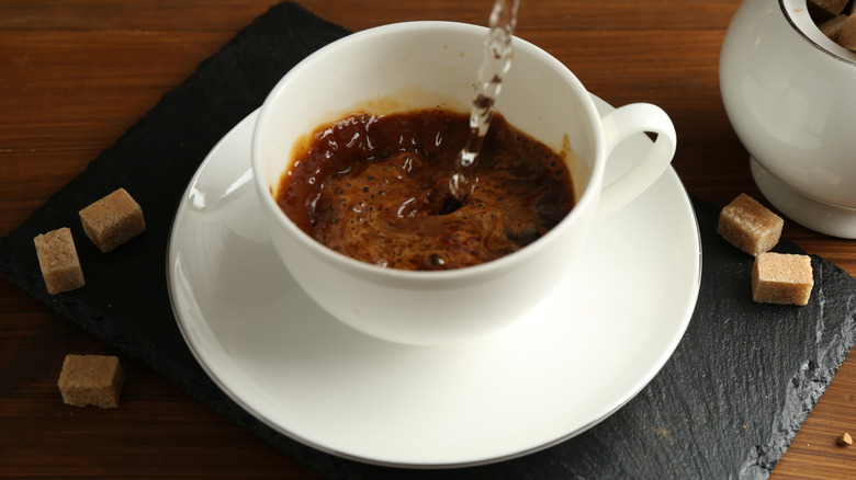 Water pouring into a white mug of instant coffee on white saucer with sugar cubes around
