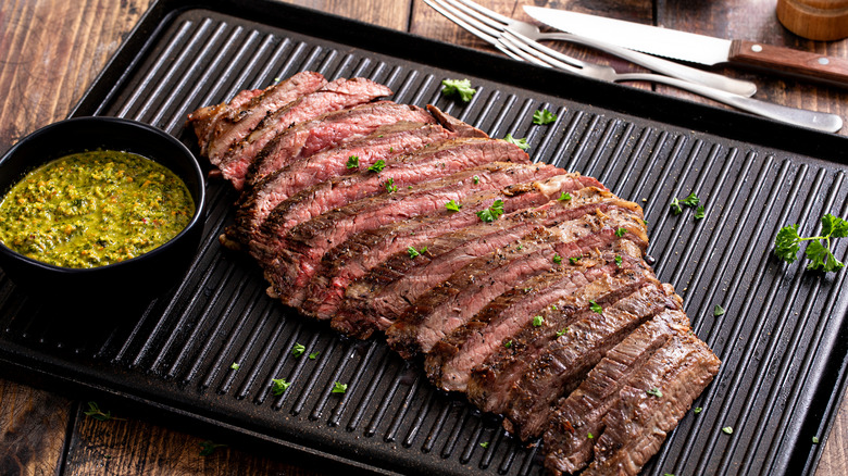 Grilled flank steak on a grill sitting next to a bowl of green sauce
