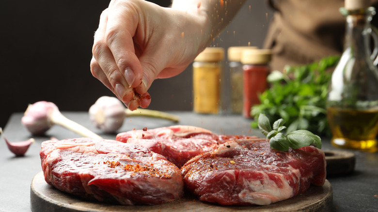 A hand sprinkling spices onto raw steak.
