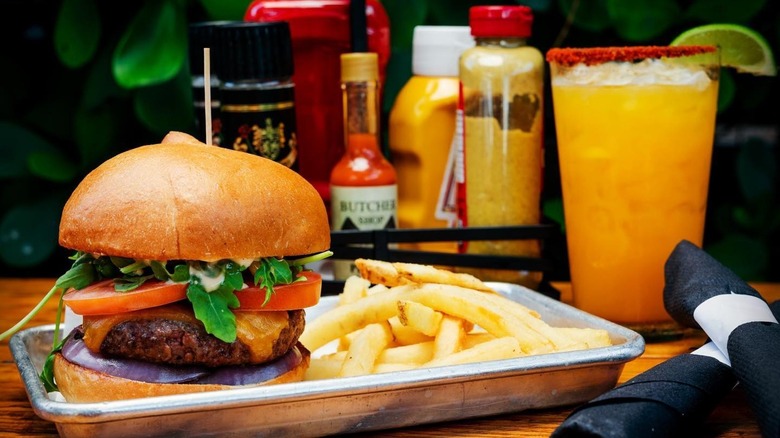 Burger and fries on a silver metal tray with condiments and a drink in the background