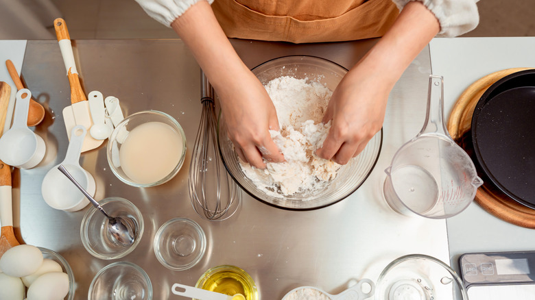 A person kneading dough in a clear bowl with baking tools around them on the table