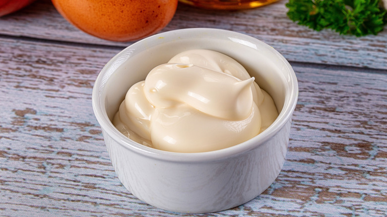 Close-up of mayonnaise in a white ramekin on a wooden surface with eggs and parsley in the background