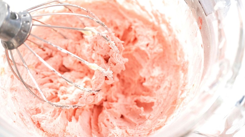 Stand-mixer metal whisk in a clear bowl of pink-hued whipped frosting