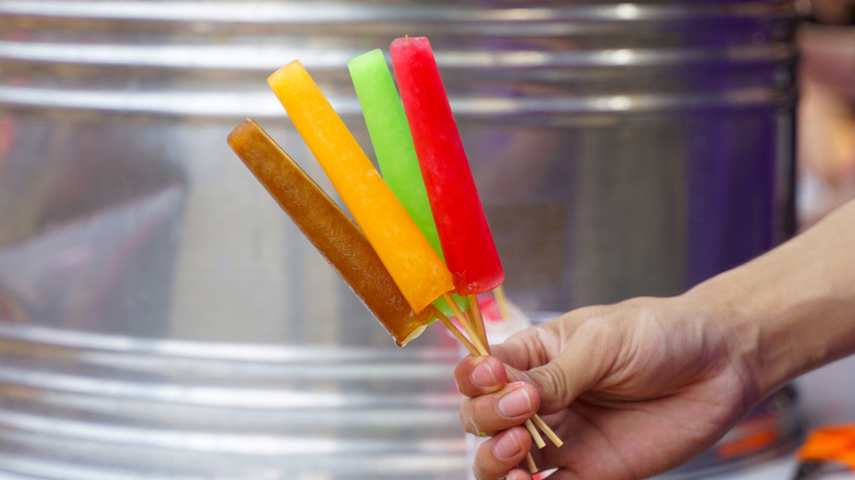 A hand holding several popsicles in front of a steel drum.