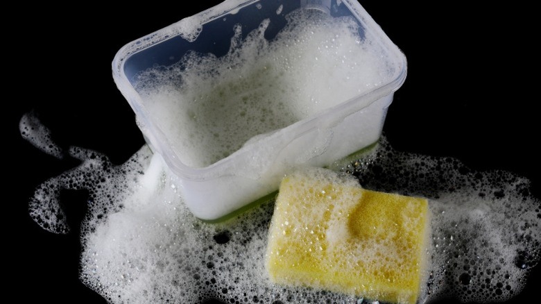 A soapy plastic food container with a cleaning sponge next to it.