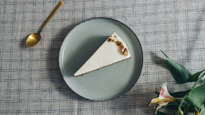 A slice of cake on a plate next to a spoon and some flowers on a checkered gray tablecloth