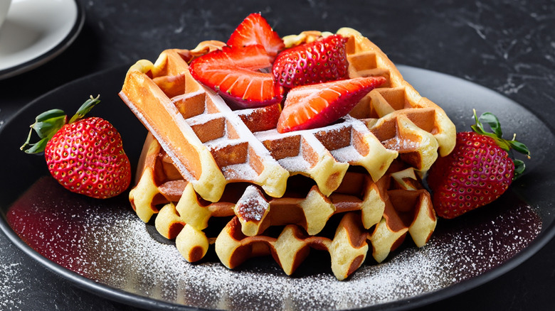 Waffles on a plate stacked with strawberries and dusted with sugar, next to a cappuccino