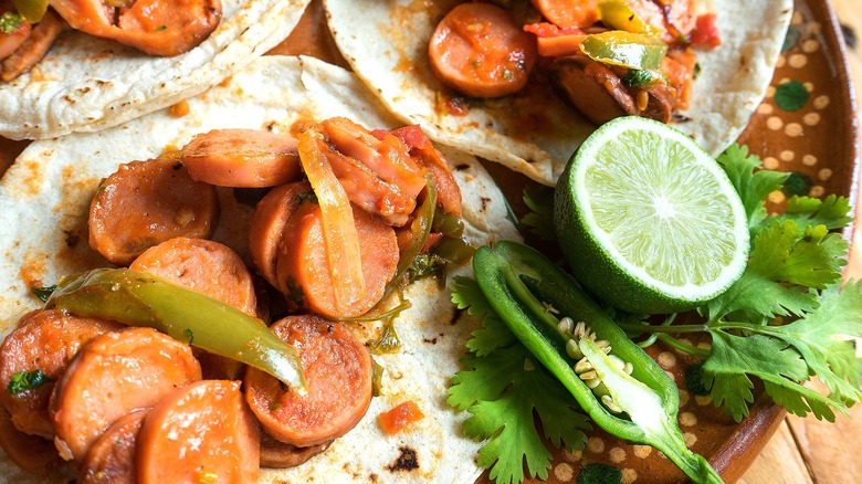 Open flour tortillas with sliced sausage and peppers on a plate with half a lime, jalapeño, and cilantro