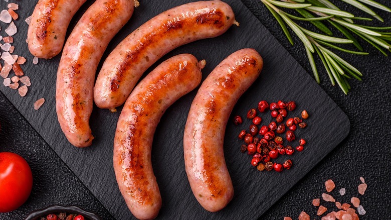 Grilled bratwurst links on a black matte serving board with coarse pink salt and whole red peppercorns