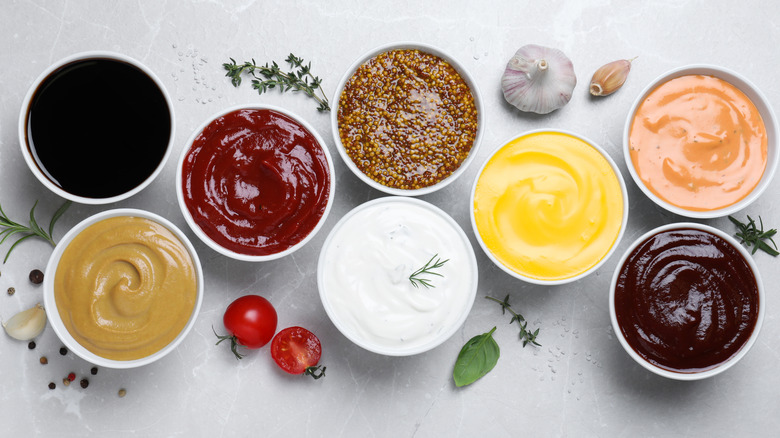 Eight bowls of various condiments on a marble table with seasoning, spices, cherry tomatoes, and garlic