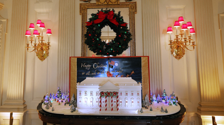 A wider shot of a gingerbread White House that looks like a realistic replica, placed on the eagle console table with miniature Christmas trees around it.