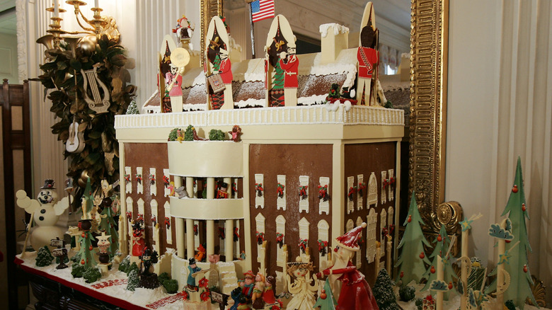 An old photo of a gingerbread White House with the American flag on top, surrounded by figurines and decorations.