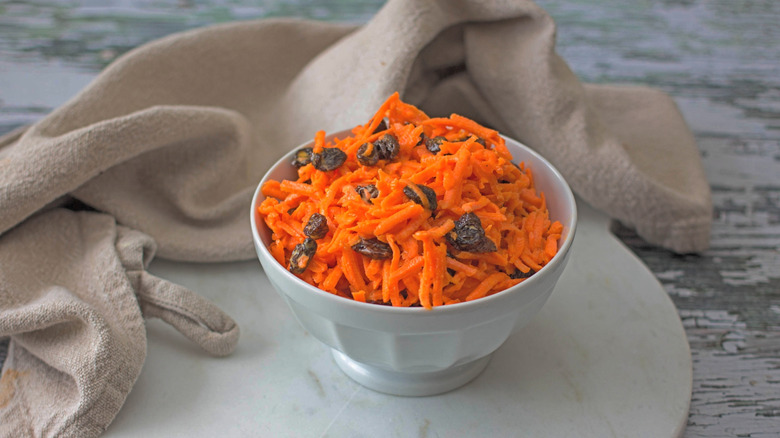A carrot and raisin salad is served on a white bowl on a round, white tray.