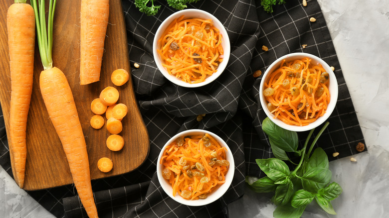 Three white bowls of carrot and raisin salad beside a cutting board topped with whole carrots.