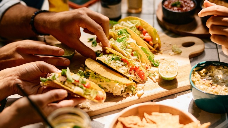 A group of people eating hard shell tacos together off of a cutting board at a table during an at-home taco night.