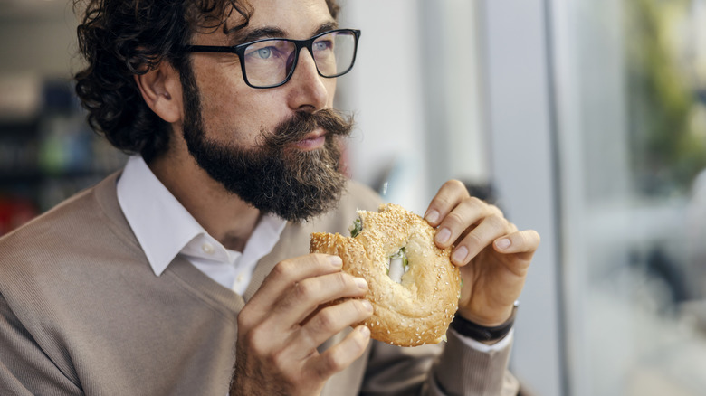 Man eating bagel sandwich and looking out a window