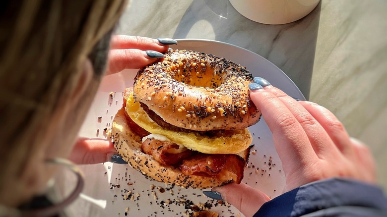View over the shoulder of person holding egg, cheese, and bacon bagel sandwich