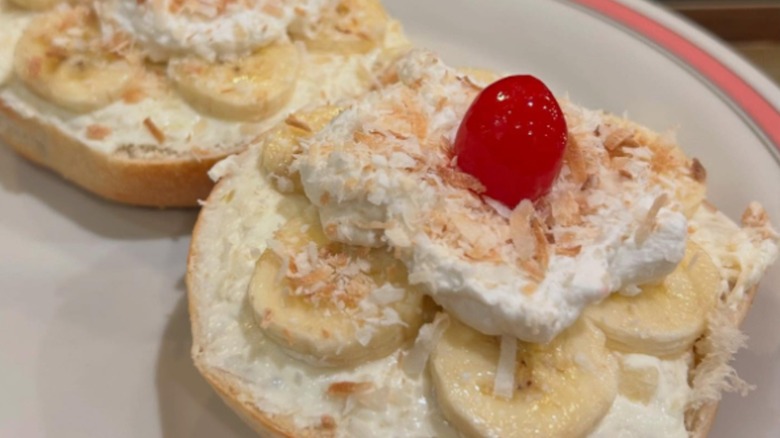 Sliced bagel with banana, cream, toasted coconut, and a cherry