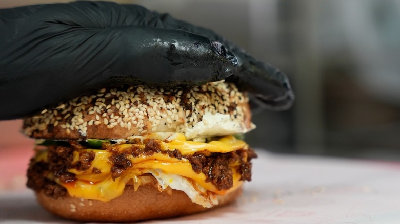 Hand with black glove pressing down on a bacon, egg, and cheese breakfast bagel