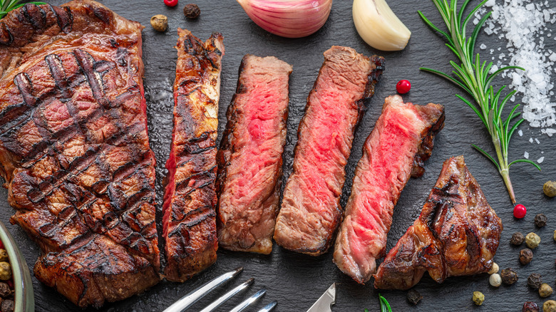 Sliced grilled ribeye on a black slate surrounded by herbs and peppercorns